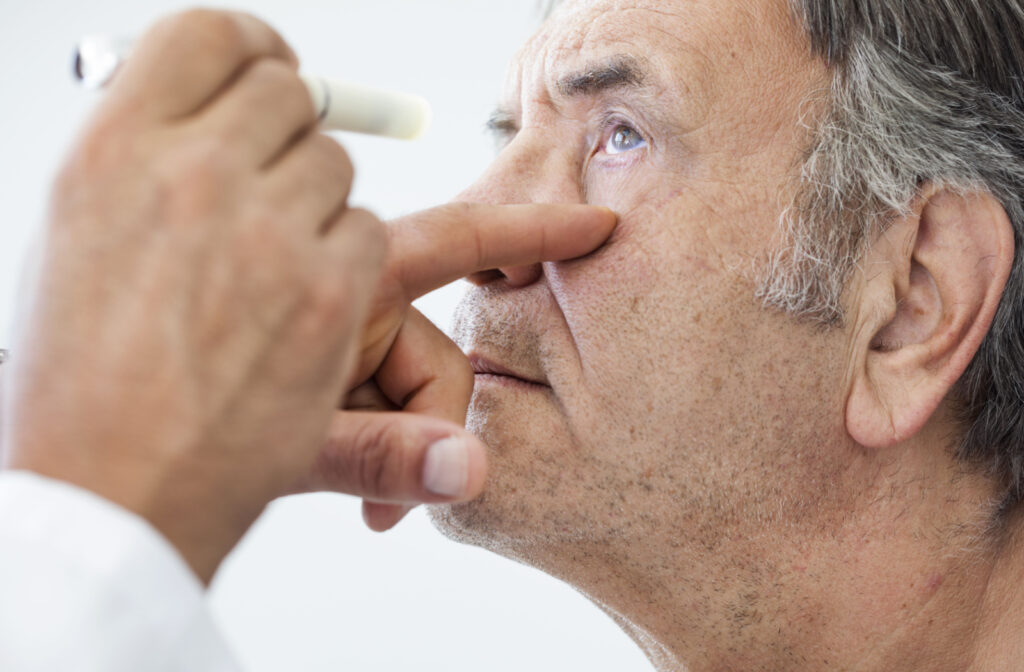 Close-up of an older man's face from the left side. He is looking up as an eye doctor shines a small light in his left eye while gently holding it open.