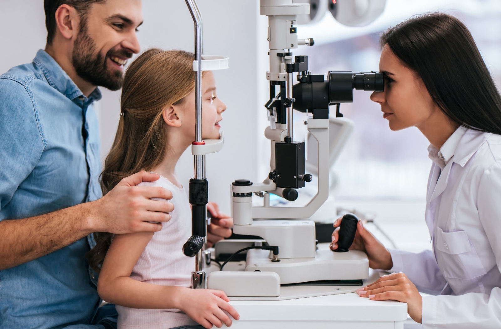 A father and daughter sit together during an exam as an optometrist examines the daughter's eyes.