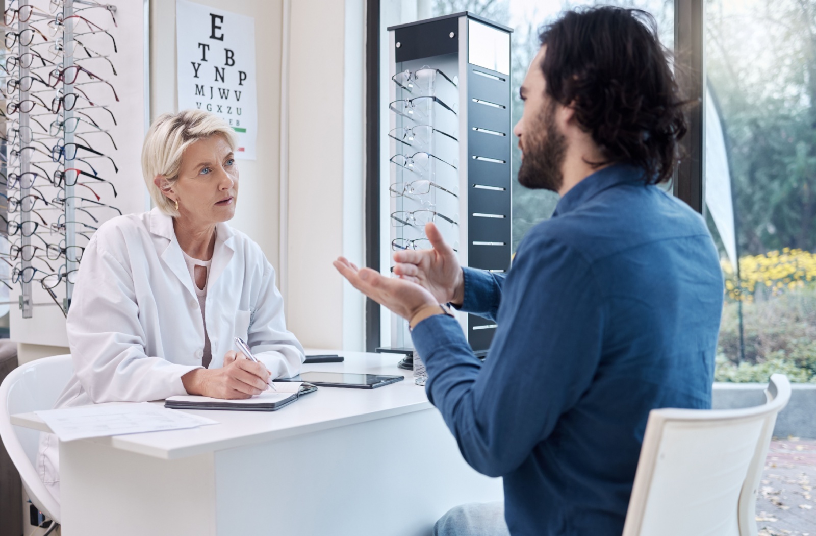 An optometrist in her office taking notes while a patient describes his recovery from laser eye surgery.