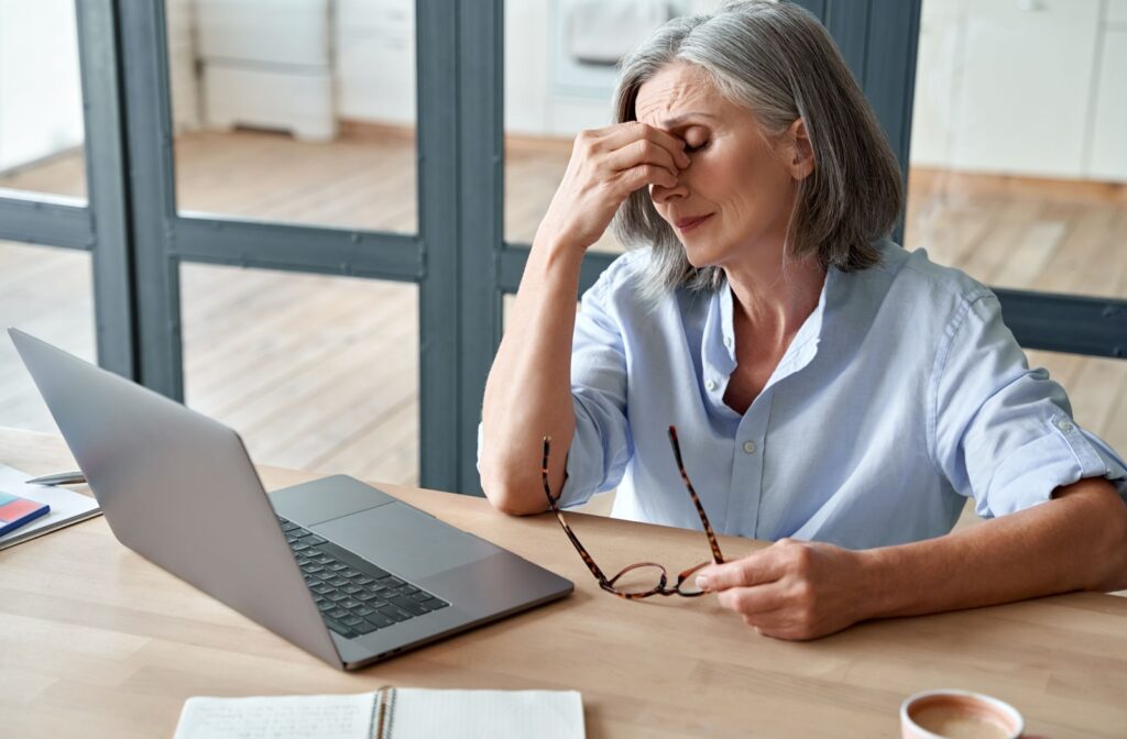 An adult taking off their glasses and rubbing their eyes in frustrating while working at their computer due to eye strain.